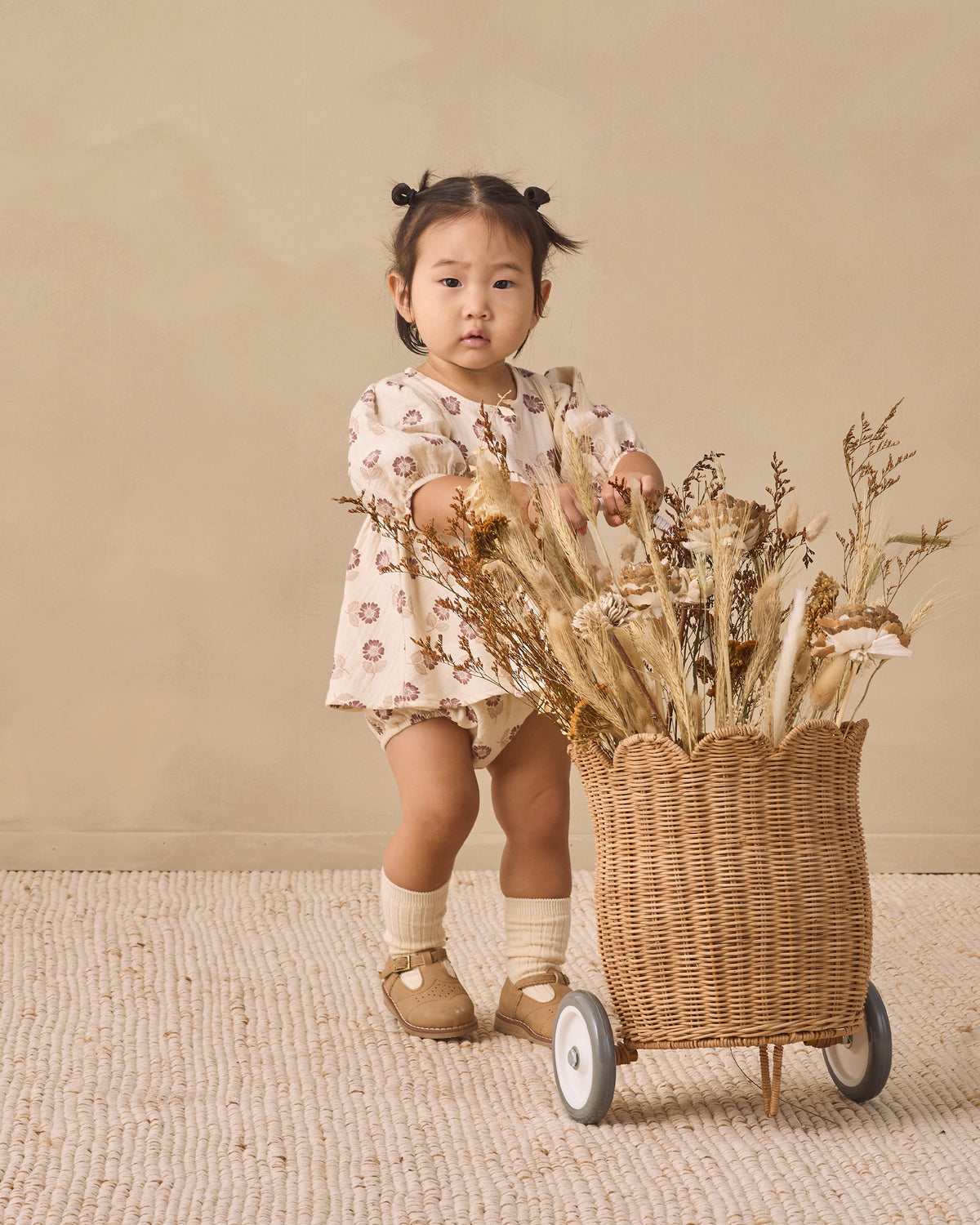 A toddler girl stands behind a wicker cart filled with dried flowers in a studio setting. She is wearing a cream-colored romper with a brown floral pattern, cream socks, and tan shoes, against a warm beige background.